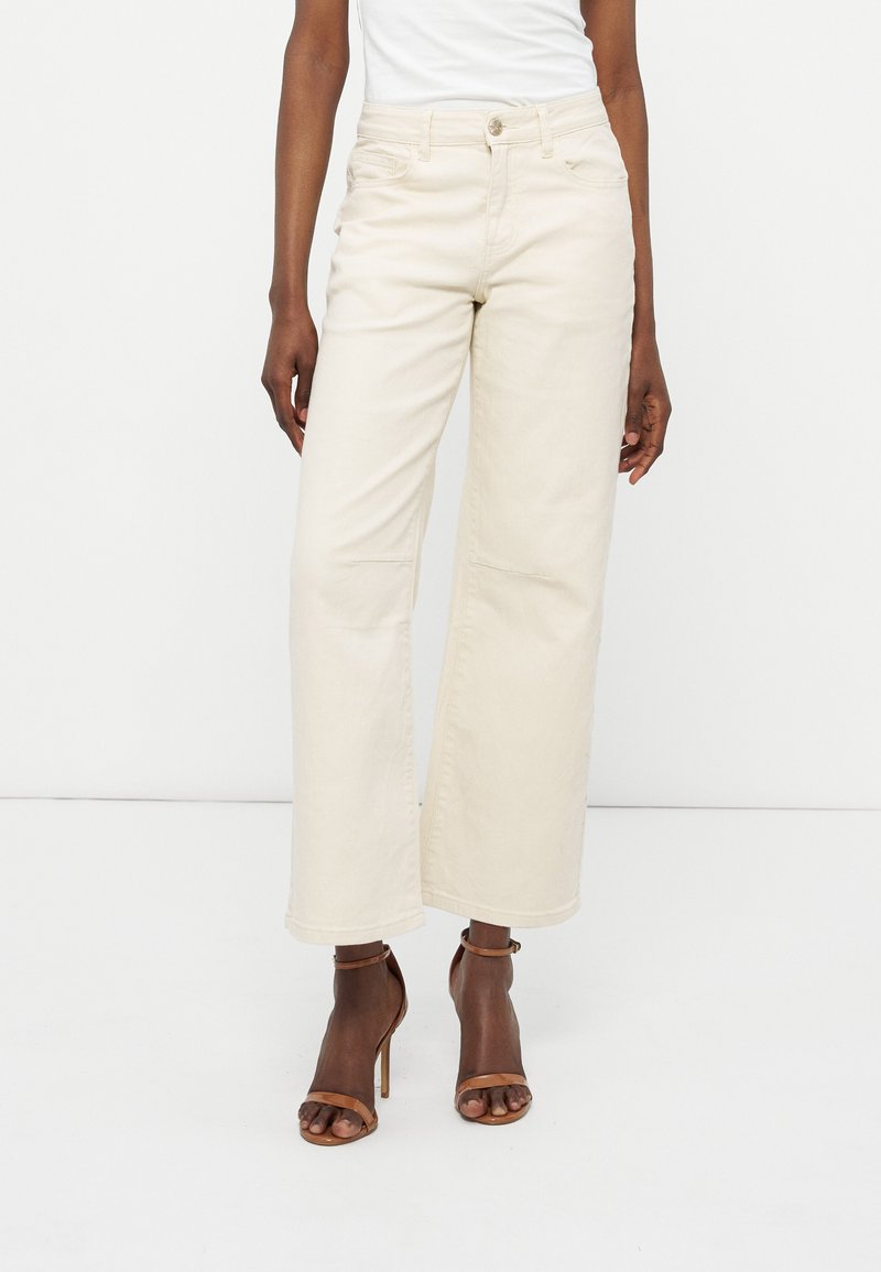 Mujer con pantalones cremosos de tiro alto y pierna ancha, y sandalias marrones de tacón alto, de pie contra un fondo blanco liso.