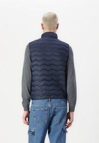 Gilet matelassé bleu marine avec un design en quilt, un col haut et un tissu léger. Porté sur une chemise à manches longues grise et un jean bleu.