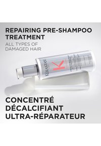 Vit cylindrisk flaska med pump, märkt "Kérastase." Följd av ett genomskinligt stativ. Grå bakgrund. Text i fetstil, svart och röd.