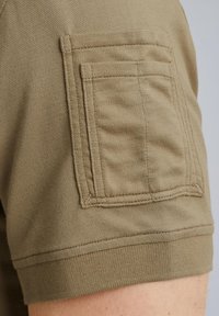 Olivengroene katoenen t-shirt met een patch zak op de linkermouw. De stof heeft een gladde textuur met een geribde manchette en onderrand.