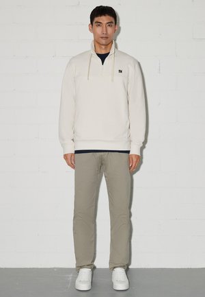 Mann steht vor einer weißen Ziegelwand und trägt einen cremefarbenen Pullover mit Viertelreißverschluss, beige Hose und weiße Turnschuhe, schaut geradeaus mit neutralem Gesichtsausdruck.