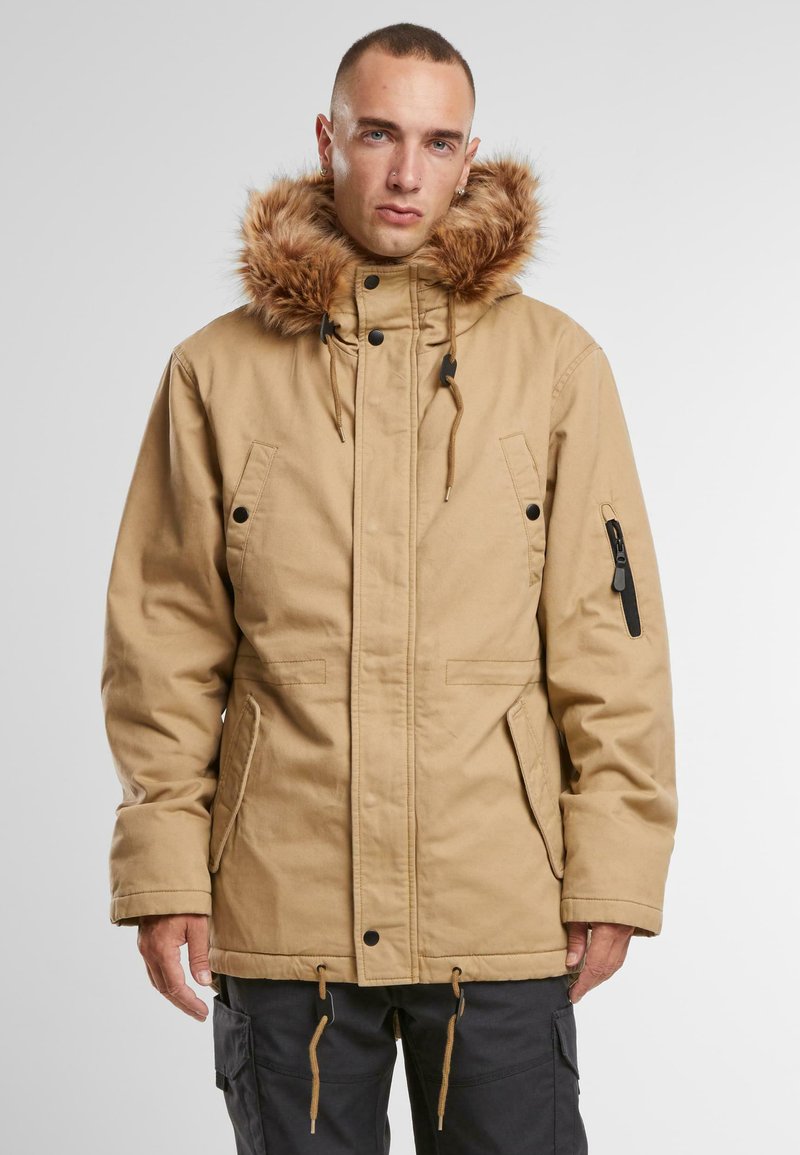 Beige parka-jakke med faux fur hette, justerbar snor, frontlommer og sideglidelåsdetalj. Tykt stoff med en glatt tekstur.