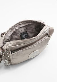 Light beige zippered crossbody bag made from smooth synthetic material. Features a prominent logo patch and a small charm accessory.