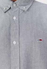 Grå knappad skjorta med en strukturerad tyg, som har en klassisk krage och vita knappar, samt en liten logotyp med blå, vita och röda accenter.