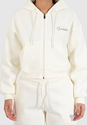 Žena, která má na sobě krémovou mikinu na zip a odpovídající kalhoty se šňůrkami, s malým textem na hrudi a motivem srdce na stehně.