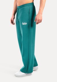 Teal joggings met een losse pasvorm, voorzien van zijzakken en een elastische tailleband. Inclusief witte "Smilodox" logo-detail aan de voorkant.