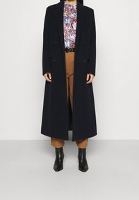 Cappotto lungo di un blu navy scuro con design doppiopetto, realizzato in un tessuto testurizzato. Il cappotto presenta un taglio classico e tasche laterali.