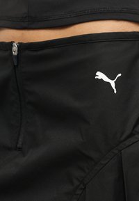 Κοντινό πλάνο μαύρου αθλητικού παντελονιού με μερικό φερμουάρ και λευκό λογότυπο Puma κοντά στη μέση.