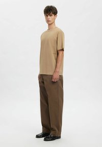 Beežikas lühikeste varrukatega T-särk, lõdva istuvusega, ühendas pruuni laiade säärtega pükstega ja mustade jalanõudega, seisab neutraalsel taustal.