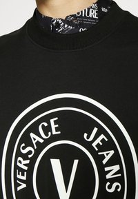 Svart sweatshirt med rund vit logotyp, "VERSACE JEANS", med ribbad halsringning och en lager-på-lager krage med grafisk tryckdetalj.