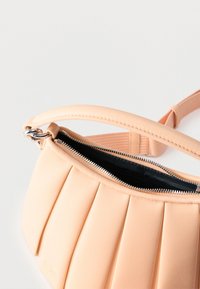 Τσάντα χειρός από δέρμα σε ανοιχτό ροδακινί χρώμα με πιέτες, ασημένιο φερμουάρ ημίκλειστο και ρυθμιζόμενο λουράκι στο ίδιο χρώμα, σε λευκό φόντο.