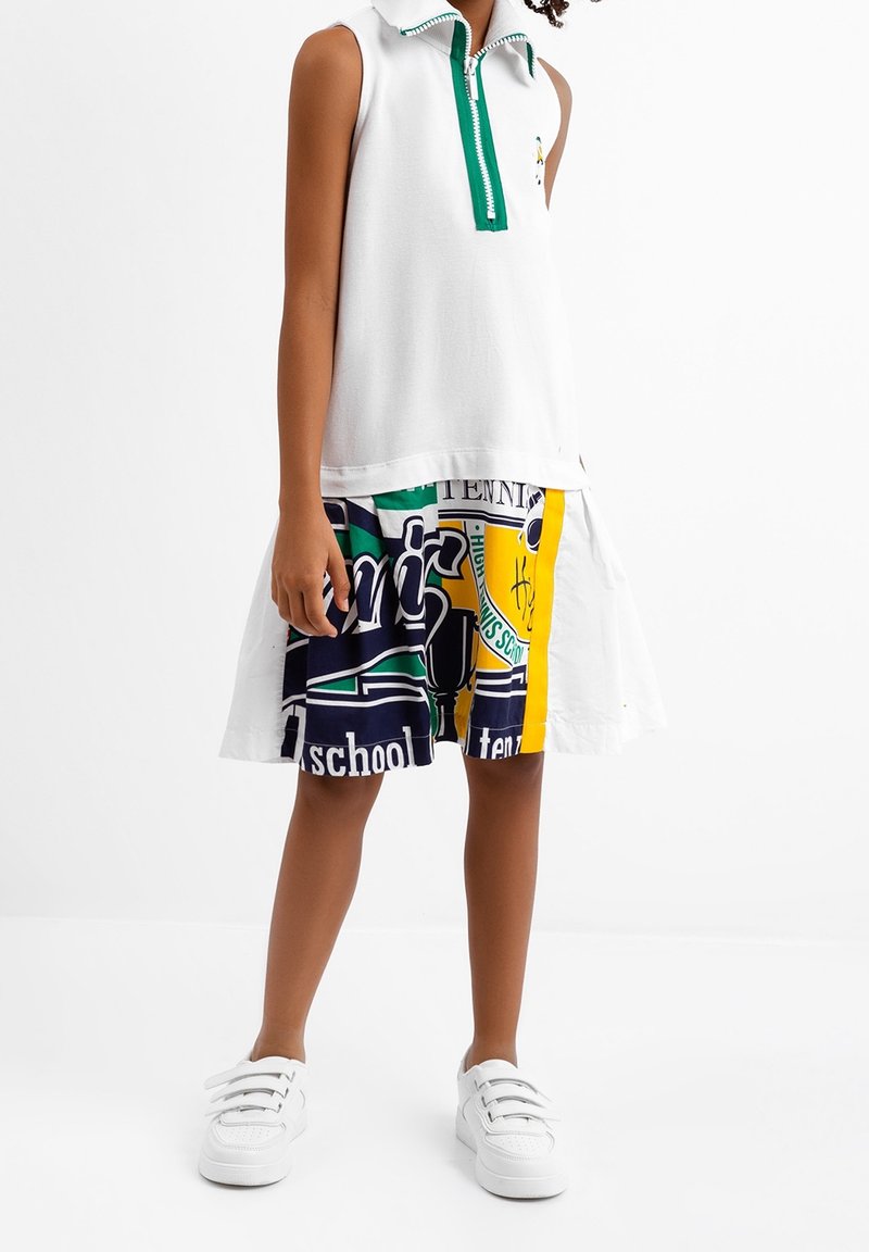 Enfant portant un haut blanc sans manches avec une fermeture éclair verte et un short graphique coloré avec des baskets blanches, debout devant un fond blanc.