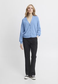 Hele sinine kootud kardigan sügava V-kaelusega, kuue nööbiga ja tekstuurse mustriga, koos mustade sirgete pükstega ja spordijalatsitega.