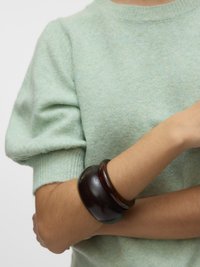 Osoba odjevena u svijetlozeleni kratki džemper s nabranim rukavima i dva široka tamnosmeđa narukvice na prekriženim rukama.