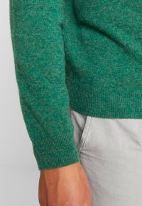 Grüner, strukturierter Strickpullover mit einem enganliegenden gerippten Bündchen. Das Material wirkt weich und leicht flauschig und zeigt einen Farbverlauf.