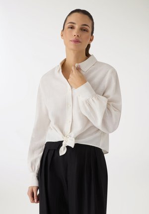 Donna con capelli scuri raccolti, indossa una camicia bianca a maniche lunghe con bottoni, annodata in vita, e pantaloni neri a vita alta su sfondo bianco.