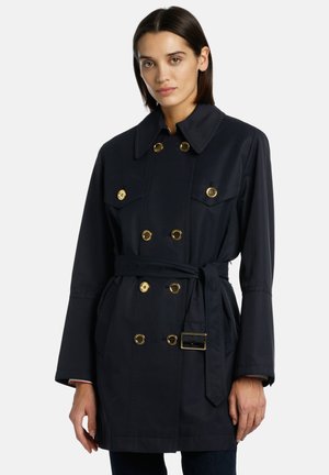 Laivastonsininen trench-coat vyötäröllä, jossa on solki, kaksirivinen muotoilu, kultaiset yksityiskohdat ja kaksi etutaskua. Sisältää suuren kauluksen ja pitkät hihat.