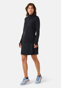 Musta urheilullinen vetoketjullinen takki korkealla kauluksella, yhdistettynä tummaan hameeseen. Jalassa vaaleansiniset juoksukengät punaisilla yksityiskohdilla.