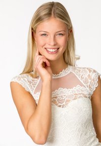 Weißes Spitzenkleid mit durchsichtiger Ausschnitt und gemustertem Oberteil. Hat kurze Spitzenärmel und eine figurbetonende Silhouette. Das Model lächelt.