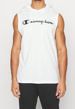 Personne musclée portant un sweat à capuche blanc sans manches avec le logo « Champion » sur la poitrine et un pantalon noir, debout devant un fond uni.
