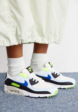 Person wearing white and blue Nike Air Max sneakers with neon green accents, white socks, and a white skirt standing on carpeted floor.