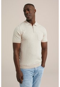 Beige short-sleeve polo shirt with a zipper collar, featuring a chest pocket and ribbed hem, paired with light blue jeans.