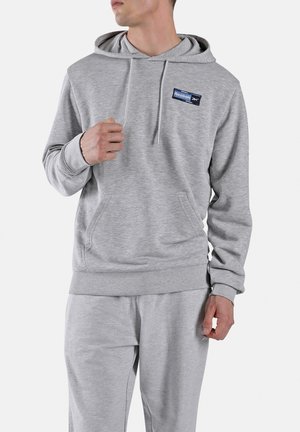 Homme portant un sweat à capuche Reebok gris clair et un pantalon de survêtement assorti, de profil, avec la main gauche près de la poitrine et la main droite détendue.