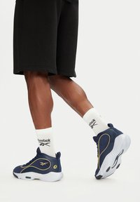 Námornícky modré športové tenisky s bielymi podrážkami, zlatými akcentmi a zelenými detailmi, skombinované s bielymi ponožkami s čiernym logom Reebok a čiernymi šortkami.