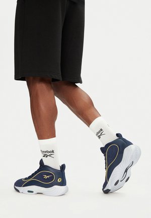 Jūras zilas sporta kurpes ar baltām zoleēm, zelta akcentiem un zaļiem detaļām, saskaņotas ar baltām zeķēm, uz kurām ir melns Reebok logo, un melniem šortiem.