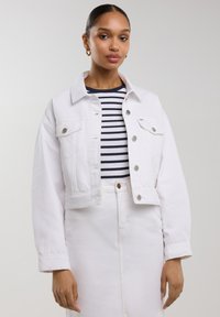 Valkoinen lyhennetty denim-takki, jossa on hopeiset napit, klassinen kaulus ja kaksi läppätaskua. Alle on pukeutunut tummansininen ja valkoinen raitapaita.