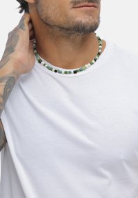 Perlekæde med grønne, sorte og hvide sten, med en glat tekstur og rund form, båret over en hvid t-shirt.