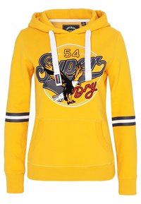 Sudadera amarilla con cuerdas blancas, gráfico de águila en blanco y negro, texto "Super Dry 54" y rayas negras en las mangas. Bolsillo tipo canguro en el frente.