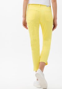 Pantalones capri amarillos hechos de tela elástica, con un diseño ajustado y bolsillos traseros, combinados con zapatillas blancas y una pequeña pulsera plateada.