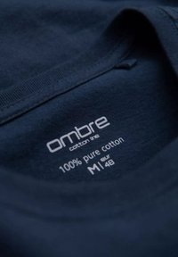 Etichetta della maglietta di cotone blu navy recita "ombre cotton line 100% cotone puro M 48." Il testo è stampato in bianco su un tessuto liscio.