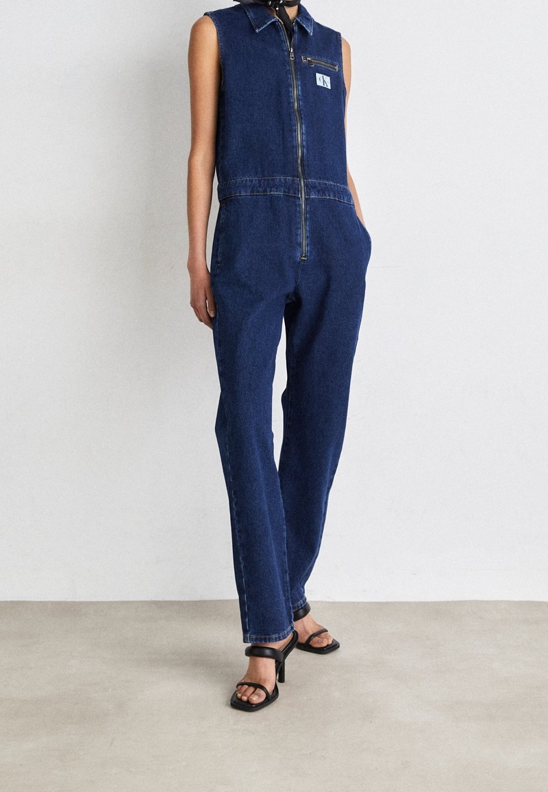 Combinaison en denim bleu foncé sans manches avec fermeture éclair devant, poche poitrine et poches latérales, portée avec des sandales à talons noires.