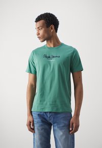 Zöld pamut póló rövid ujjakkal, kerek nyakkivágással, és sötétkék "Pepe Jeans London" felirattal a elején. Világoskék farmernadrággal viselve.