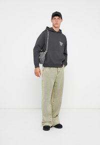 Šedá mikina s přední kapsou, světle zelené široké džíny, černé sandály a šedá crossbody taška. Model má na hlavě černou kšiltovku.