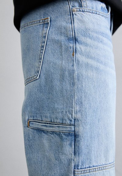 Jeans din denim de culoare albastru deschis, cu un design drept, având cusături vizibile la buzunare și o textură subtilă. Două buzunare laterale sunt vizibile.