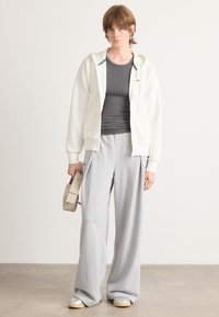 Bijela zip-up hoodie, sivi uski top, sivi široki trenirci i sivi tenisice. Model drži malu bež torbu. Jednostavan dizajn.