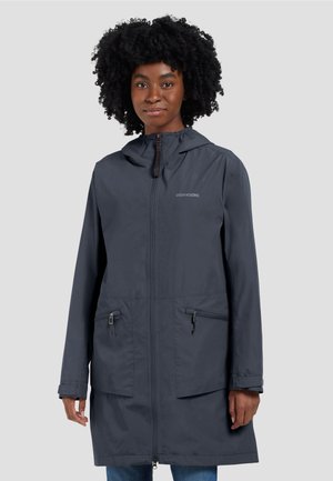 Woman wearing a navy blue hooded raincoat with front zipper and two zippered pockets, standing against a plain light gray background.