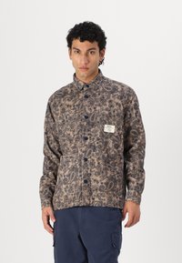 Beige hosszú ujjú ing sötétkék paisley mintával, gombos elülső rész, és egy címkés mellzseb. Sima szövet textúra.