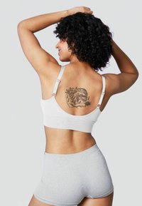 Reggiseno sportivo bianco con spalline regolabili, caratterizzato da una texture liscia. Pantaloni corti grigi a vita alta. Design del tatuaggio visibile sulla schiena.