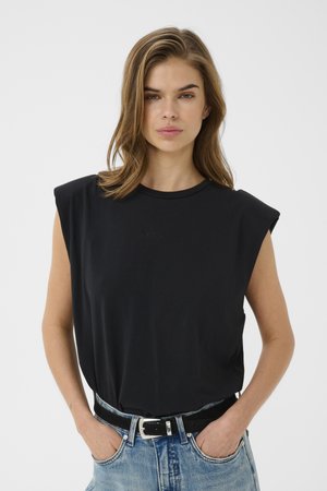 Junge Frau mit schulterlangem braunen Haar, die ein schwarzes ärmelloses Top und blaue Jeans trägt, Hände in den Taschen, neutraler Gesichtsausdruck.