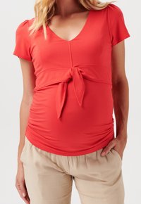 Top rosso a maniche corte con scollatura a V, dettaglio con nodo frontale e lati arricciati, abbinato a pantaloni beige larghi. Tessuto morbido.