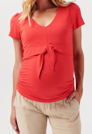 Top rosso a maniche corte con scollatura a V, dettaglio con nodo frontale e lati arricciati, abbinato a pantaloni beige larghi. Tessuto morbido.