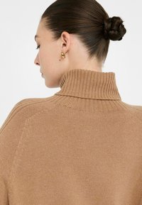 Donna con i capelli raccolti in un chignon intrecciato, che indossa un maglione a collo alto beige e orecchini a cerchio dorati, voltata di lato di fronte a uno sfondo semplice.