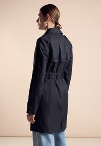 Ciemnogranatowy trencz z szerokim kołnierzem i paskiem w talii. Posiada wentylację z tyłu oraz mankiety zapinane na guziki. Stylizowany z jasnoniebieskimi spodniami.