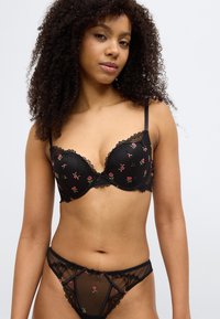 Σετ μαύρου εσωρούχου που περιλαμβάνει σουτιέν με επένδυση και κεντήματα λουλουδιών σε ροζ και πράσινο, συνοδευόμενο από διαφανές δαντελένιο string σετ.