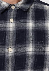 Námořnická a bílá kostkovaná flanelová košile s knoflíkovým límečkem, která má krémové knoflíky a vystouplé logo na látce.