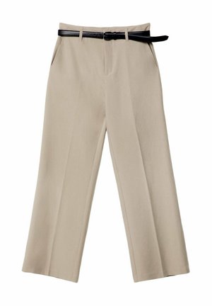 Persoon die beige broek met wijde pijpen, een zwarte riem, een navy ritstop met een zwarte trui over de schouders gedrapeerd en zwarte schoenen draagt, staand tegen een witte muur.